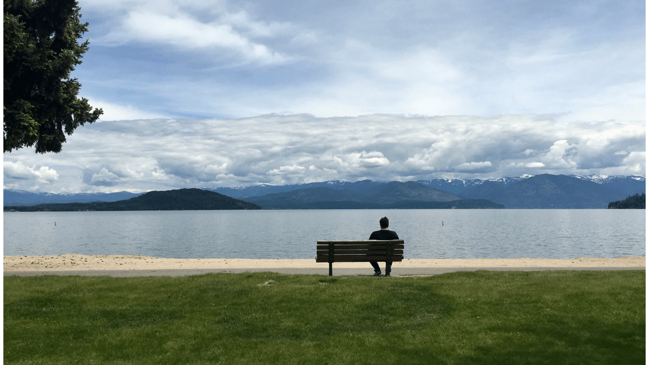 A person sits on a park bench in front of a large lake and mountains